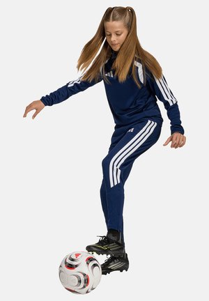 Jeune fille en tenue de sport bleu marine à bandes blanches contrôlant un ballon de football avec son pied sur un fond clair et uni.