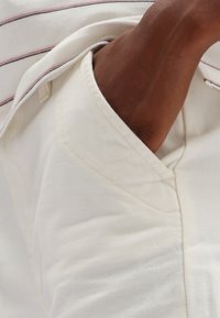 Vit tyg med en texturerad yta, som har en fickkant och en glimt av en brun arm. Randig detaljering i röd och svart synlig.