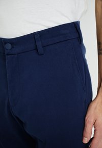 Pantalons ajustés bleu marine avec une texture lisse, devant plat, poches latérales et fermeture à bouton à la taille. Design classique, coupe slim.