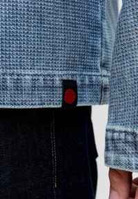 Tissu bleu texturé d'une veste en denim avec un accent circulaire rouge sur une étiquette noire au bas, associé à un pantalon en denim foncé.