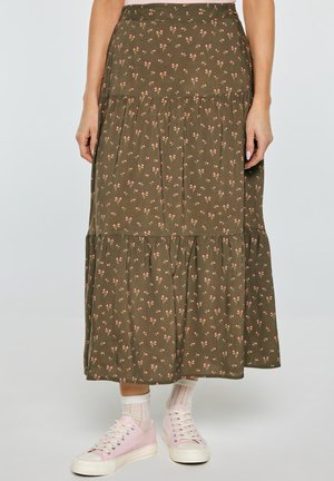 Kobieta ubrana w oliwkowo-zieloną midi spódnicę w kwiaty w kolorze różowym i białym, do tego białe skarpetki i jasnoróżowe trampki.