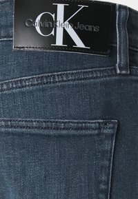 Jeans de mezclilla en azul oscuro con un acabado texturizado, que presentan una etiqueta de cuero negro con "Calvin Klein Jeans" en texto blanco y detalles de costura visibles.