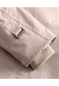Manica di giacca rosa chiarissimo con una superficie liscia, polsino a maglia beige in contrasto e un accento in metallo su un'etichetta di tessuto.