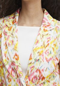 Blommig kavaj med en blandning av gula, rosa och bruna mönster på en vit bakgrund, med spetsig krage och lätt tyg.