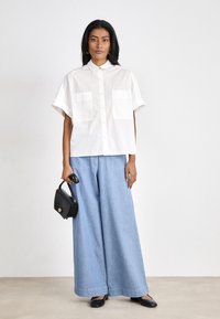 White short-sleeve shirt with two front pockets, paired with light blue wide-leg trousers. Holding a small black handbag, wearing black shoes.