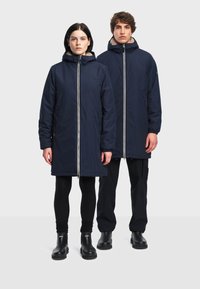 Marineblauwe lange jassen met een ritssluiting aan de voorkant, een capuchon en fleece voering. Beiden dragen zwarte broeken en laarzen, wat een eenvoudig, functioneel ontwerp laat zien.