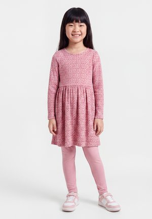 Ung pige smiler, iført en lyserød langærmet kjole med små blomsterprint, lyserøde leggings og hvide og lyserøde sneakers, står på en hvid baggrund.