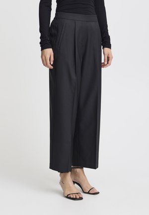 Pantaloni neri a gamba larga con una texture liscia, dotati di vita alta, pieghe e tasche laterali. Abbinati a sandali neri sottili.