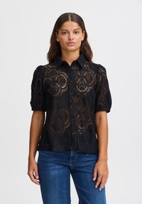 Blusa nera di pizzo con motivi floreali a taglio, maniche a sbuffo corte, chiusura con bottoni sul davanti e colletto. Indossata con jeans blu.
