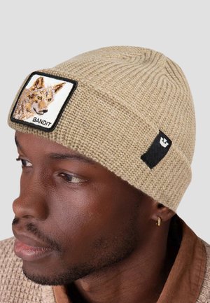 Uomo con un berretto lavorato a maglia beige con un patch a forma di testa di volpe e la parola "BANDIT" sul davanti, un piccolo etichetta nera sul lato e un orecchino a cerchio dorato.