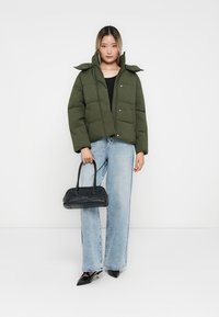 Chaqueta acolchada de color verde oliva con botones a presión y capucha, llevada sobre una camiseta negra y unos jeans de pierna ancha de color azul claro. Bolso negro y zapatos de punta.