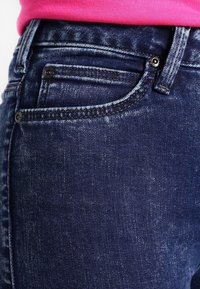 Jeans de mezclilla azul oscuro con un diseño clásico de cinco bolsillos, un pequeño bolsillo en la parte delantera y detalles de costura visibles.