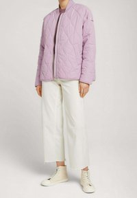 Gepatchte lila Jacke mit einem Reißverschluss vorne, geripptem Kragen und langen Ärmeln. Kombiniert mit weißen weit geschnittenen Jeans und beigen High-Top-Sneakern.