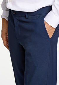 Granatowe spodnie wizytowe wykonane z teksturowanej tkaniny, z przednim płaskim krojem, bocznymi kieszeniami oraz zapinane na guzik w talii.