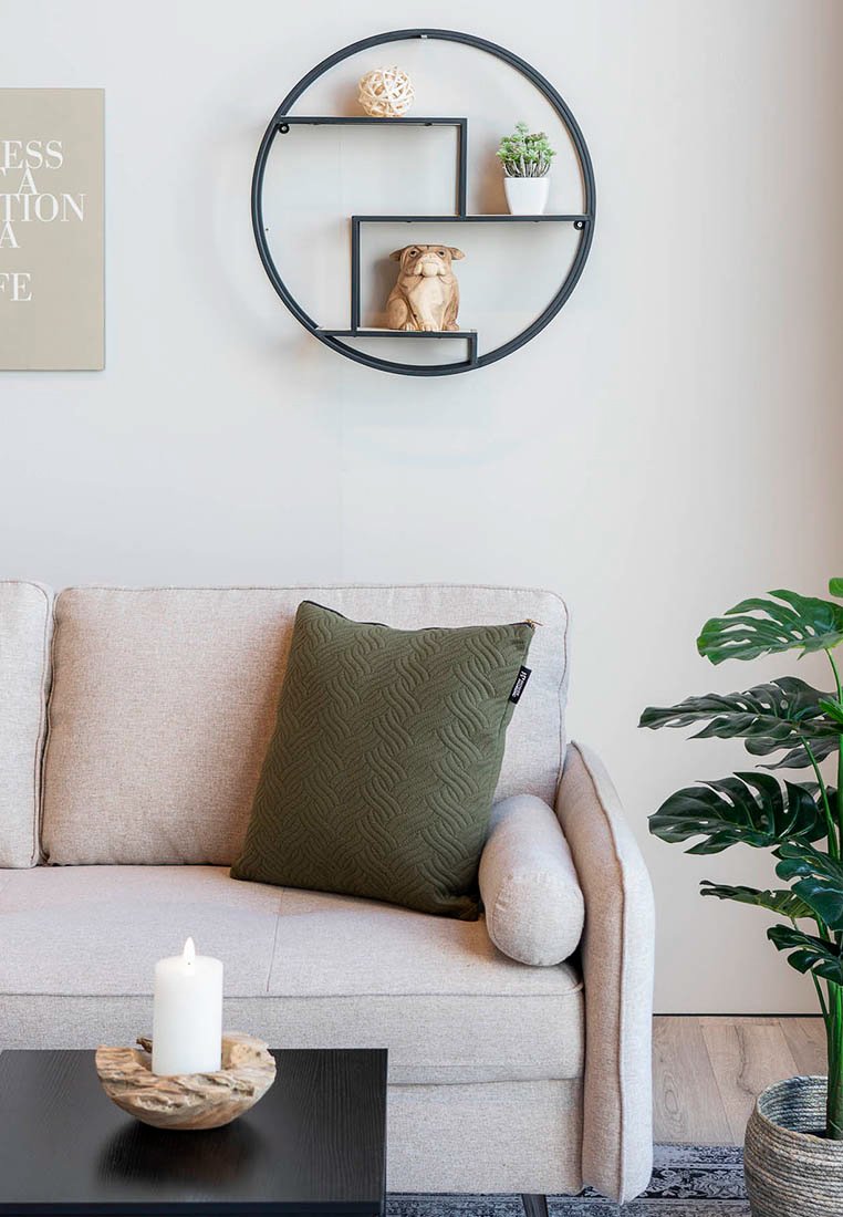 Beżowa sofa z zieloną poduszką, okrągła czarna półka ścienna z rośliną, rzeźbą, zapaloną białą świecą na czarnym stoliku kawowym oraz zielona roślinność.