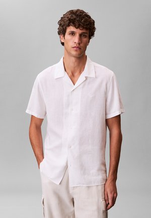 Junger Mann mit lockigem Haar, der ein weißes Hemd mit kurzen Ärmeln und beige Hosen trägt, steht vor einem schlichten grauen Hintergrund.