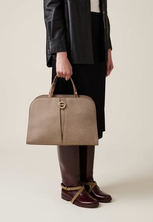 Braune Handtasche mit strukturierter Oberfläche und abgerundetem oberen Teil, Ledergriffen und goldener, runder Akzent. Kombiniert mit einem schwarzen Rock und burgunderfarbenen Stiefeln.