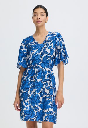 Kobieta ubrana w białą sukienkę do kolan z dużym niebieskim kwiatowym nadrukiem, z krótkimi, falującymi rękawami, dekoltem w serek i paskiem do wiązania w talii na jednolitym tle.