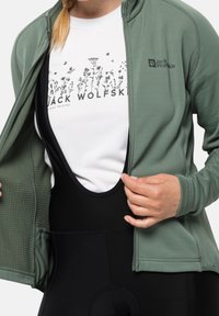 Giacca verde testurizzata aperta sopra una t-shirt bianca con stampa floreale e pantaloni da bib neri. Presenta un logo sul petto della giacca.