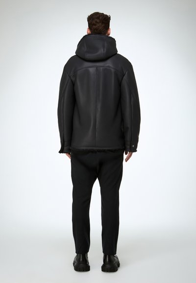 Schwarze Lederjacke mit Kapuze, mit glatter Oberfläche, Fransen am Saum und großzügiger Passform. Kombiniert mit schwarzer Hose und klobigen Schuhen.