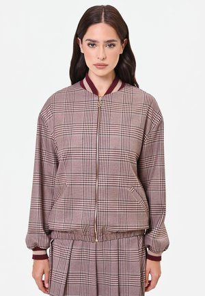 Donna con capelli scuri che indossa una giacca con zip a quadri marrone e bordeaux e una gonna plissettata abbinata su sfondo bianco.
