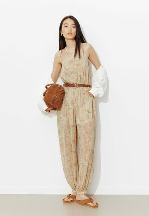 Jeune femme portant une combinaison fleurie avec une ceinture marron, une veste blanche drapée, tenant un sac en daim marron, chaussée de sandales et portant un collier de perles, debout contre un mur blanc.