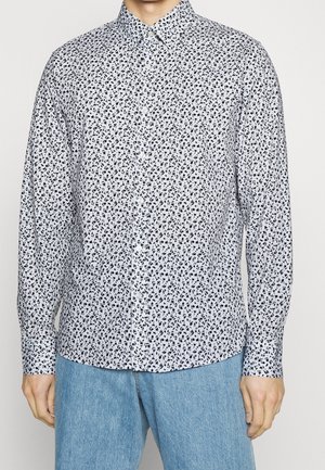 Mężczyzna w białej koszuli zapinanej na guziki z małym czarnym kwiatowym wzorem i jasnoniebieskich dżinsach, stojący na jednolitym tle.