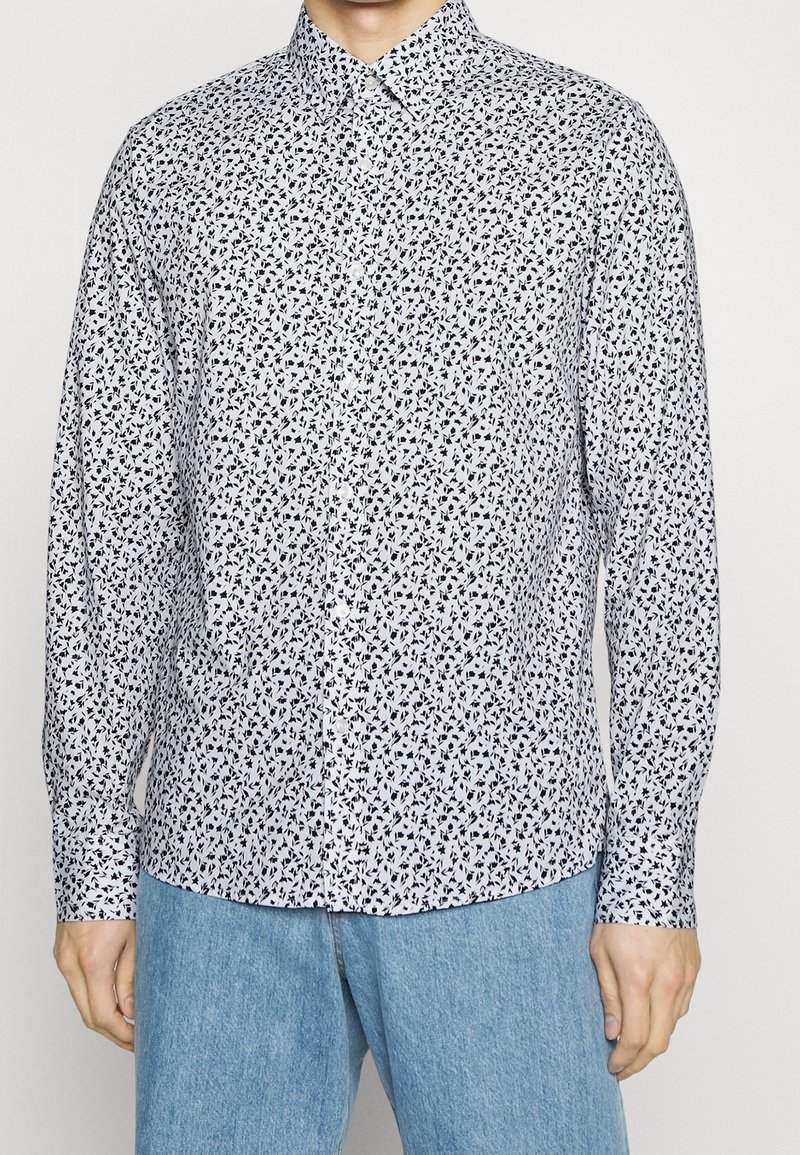 Homme portant une chemise blanche à boutons avec un petit motif floral noir et un jean bleu clair, debout devant un fond uni.