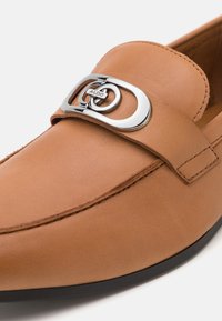 Mocassin en cuir marron avec un bout arrondi, doté d'une boucle métallique argentée avec des cercles entrelacés et des détails cousus le long de la tige.