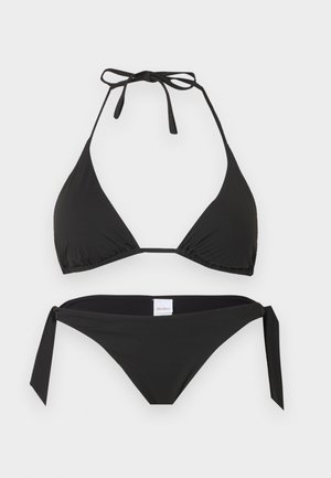 Frau, die vor einfarbigem Hintergrund steht, trägt einen schwarzen Neckholder-Bikini mit seitlich gebundenen Höschen und schwarzen Slide-Sandalen.