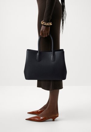 Person wearing brown ribbed dress and brown pointed low-heel shoes, holding a black leather handbag and wearing a chunky gold bracelet.