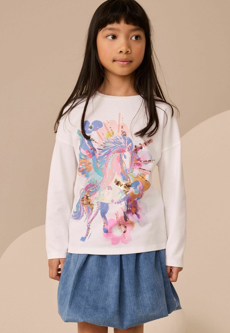 Niña joven con cabello largo y oscuro que lleva un suéter blanco con un unicornio colorido con lentejuelas y una falda vaquera azul.