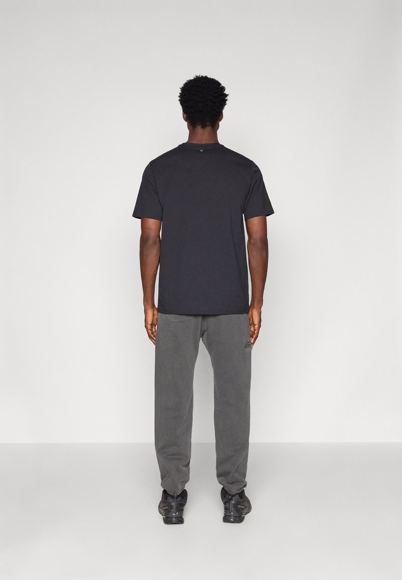 Black cotton t-shirt with a crew neck, paired with gray pants. The model stands facing away, showcasing the back of the outfit.