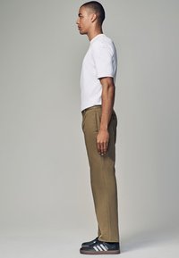 Vitt kortärmad t-shirt, khakifärgade byxor, svarta sneakers med vita accenter. Modellen visad i profil mot en neutral bakgrund.