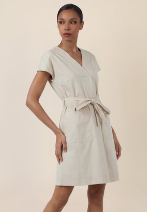 Frau trägt ein beigefarbenes Kurzarmkleid mit V-Ausschnitt und gebundenem Taillengürtel, steht vor einem schlichten neutralen Hintergrund.