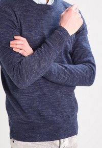 Pull en tricot bleu marine avec un motif texturé, col rond et poignets côtelés, porté par une personne aux bras croisés.