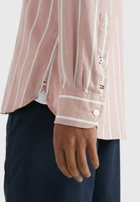 Camicia a righe rosa con colletto a punta, polsi con bottoni, righe bianche e tessuto contrastante all'orlo. Tessuto liscio e leggero.