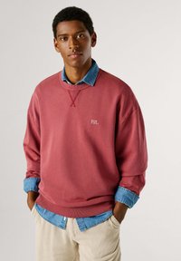 Sudadera de gran tamaño color rosa con el logo bordado "PJL", combinada con una camisa de mezclilla azul claro, llevada con pantalones de pana color crema.