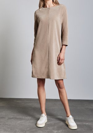 Vrouw in een beige gebreid jurk tot op de knie met driekwartmouwen en witte sneakers, staand op een grijze vloer tegen een lichtgrijze muur.