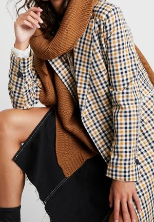 Femme portant une écharpe en tricot marron, un blazer à carreaux et une jupe noire, assise avec la main près du visage et le genou visible.