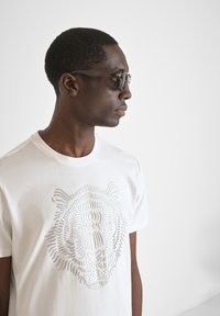 Witte katoenen T-shirt met een getextureerde leeuwafbeelding in een subtiele tint. Model draagt donkere zonnebril en kijkt iets zijwaarts.