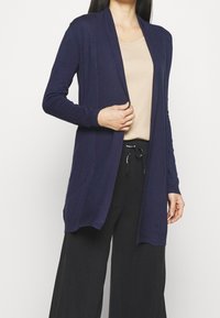 Cardigan aperto blu navy realizzato in tessuto a maglia testurizzato, con maniche lunghe e due tasche frontali, abbinato a una maglietta beige e pantaloni neri.