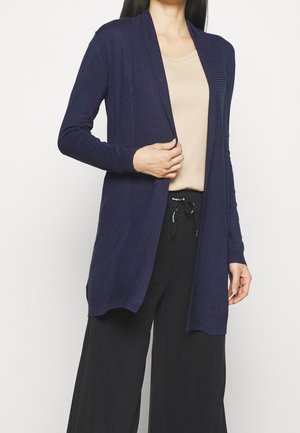 Cardigan aperto blu navy realizzato in tessuto a maglia testurizzato, con maniche lunghe e due tasche frontali, abbinato a una maglietta beige e pantaloni neri.