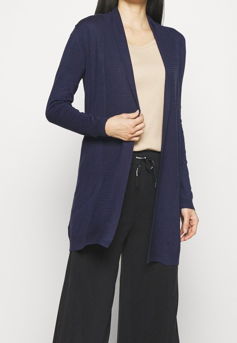 Cardigan aperto blu navy realizzato in tessuto a maglia testurizzato, con maniche lunghe e due tasche frontali, abbinato a una maglietta beige e pantaloni neri.