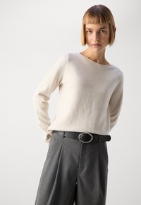 Light beige sweater with a crew neckline, paired with gray pleated trousers. A black belt with an oval silver buckle accents the outfit.