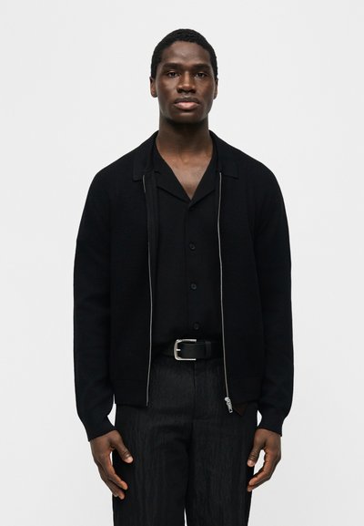 Άνδρας ντυμένος με ολομαύρο σύνολο, που περιλαμβάνει ζακέτα με φερμουάρ εμπρός, πουκάμισο με γιακά, πλισέ παντελόνι και ζώνη, που στέκεται μπροστά σε απλό ανοιχτόχρωμο φόντο.