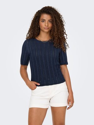 Jeune femme aux cheveux bouclés portant un top en maille bleu marine à manches courtes et un short blanc, debout avec une main dans la poche sur un fond uni.