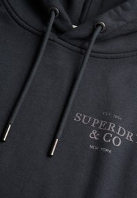 Mørkeblå hættetrøje lavet af blødt stof, med en forreste kængurulomme, justerbar snørelukning i hætten og et trykt logo i sølv.