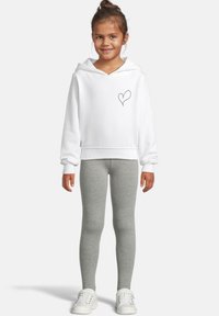Barn som har på seg en hvit hettegenser med et lite hjerte-design, grå leggings og hvite joggesko, står og smiler mot en ensfarget bakgrunn.