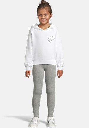 Lapsi, jolla on päällä valkoinen huppari, jossa on pieni sydänkuvio, harmaat leggingsit ja valkoiset lenkkarit, seisoo ja hymyilee yksinkertaista taustaa vasten.